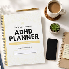 An ADHD planner notebook with a white cover and black text is shown in a styled flat-lay arrangement on a light surface, surrounded by a coffee cup, pen, phone, small plant, and another notebook.