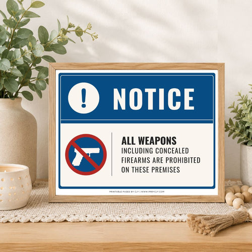 A framed warning sign displayed on a wooden table in a styled home setting. The sign features a blue and white background with the text 'NOTICE' and 'ALL WEAPONS INCLUDING CONCEALED FIREARMS ARE PROHIBITED ON THESE PREMISES' alongside a red-and-white crossed-out gun icon.