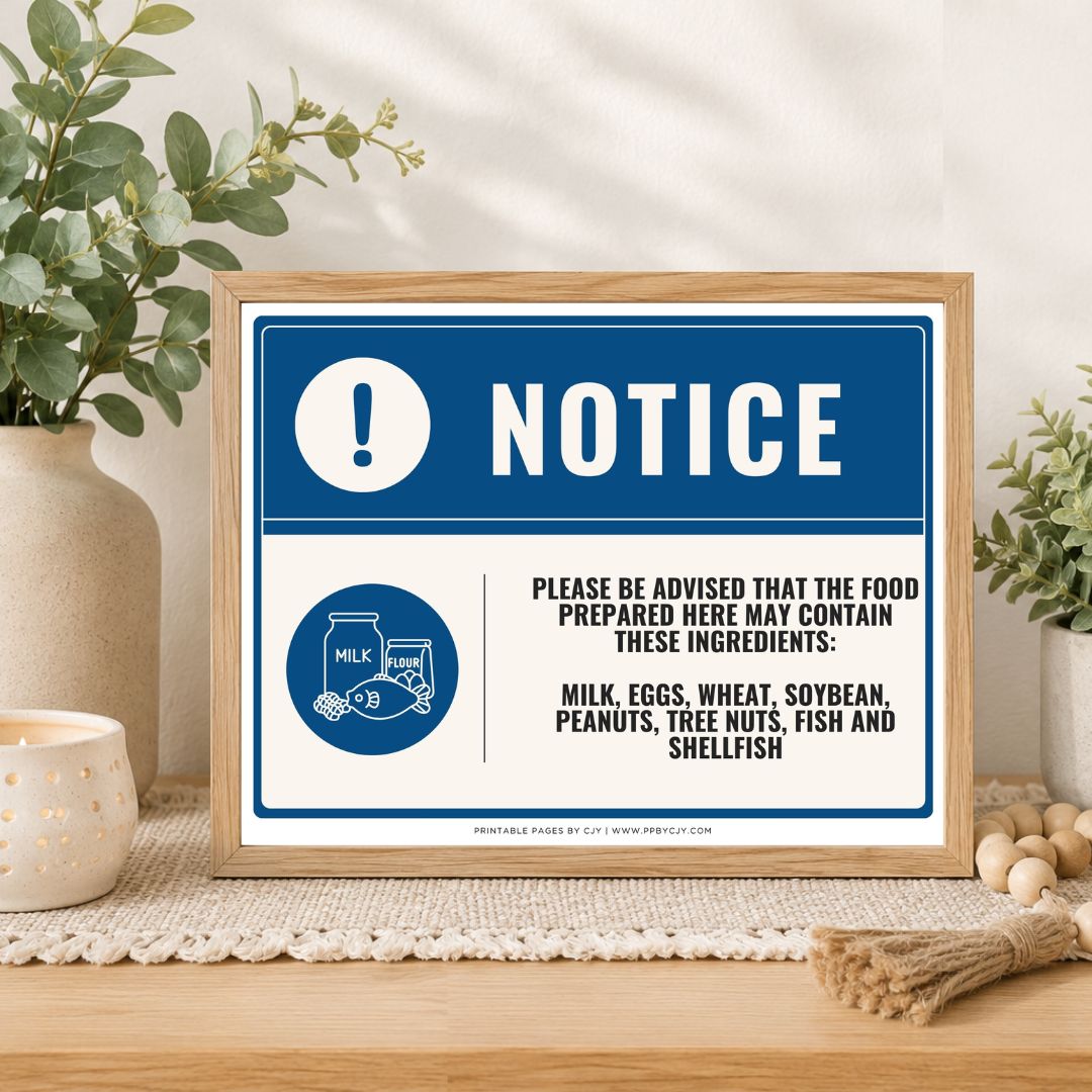 A framed food safety notice sign displayed on a wooden table in a styled kitchen or cafe setting with plants and a candle. The sign features a blue and white design with text warning about allergens including milk, eggs, wheat, soybean, peanuts, tree nuts, fish, and shellfish.