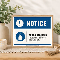 A framed printed apron-required notice sign sits on a wooden surface in a styled home setting with plants, a candle, and decorative beads. The sign has a blue and white design with text reading 'NOTICE' and 'APRON REQUIRED THANK YOU FOR YOUR COOPERATION.