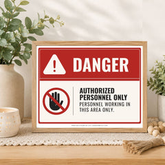 A framed warning sign with a red and white background, featuring the word 'DANGER' and a crossed-out hand icon with the text 'AUTHORIZED PERSONNEL ONLY PERSONNEL WORKING IN THIS AREA ONLY.' The sign is displayed on a wooden surface with decorative plants and candles in the background.