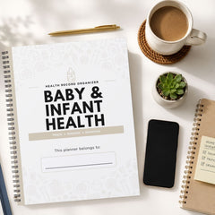 A spiral-bound baby and infant health record organizer planner lies on a white desk, surrounded by a coffee cup, a pen, a small succulent, a smartphone, and another notebook.