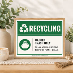 A framed recycling sign displayed on a wooden table in a styled home setting. The sign features a green and white design with the text 'RECYCLING', 'BAGGED TRASH ONLY', and 'THANK YOU FOR HELPING KEEP OUR PLANET CLEAN.