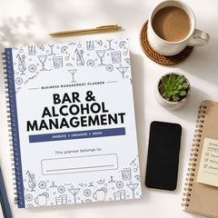 A spiral-bound planner titled 'BAR & ALCOHOL MANAGEMENT' lies on a white desk, surrounded by a cup of coffee, a smartphone, a small succulent, and another notebook.