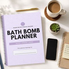A spiral-bound Bath Bomb Planner notebook lies open on a white desk, surrounded by a coffee cup, pen, small succulent, smartphone, and another notebook. The planner cover is light lavender with black text reading 'BATH BOMB PLANNER' and a purple circular logo near the top.