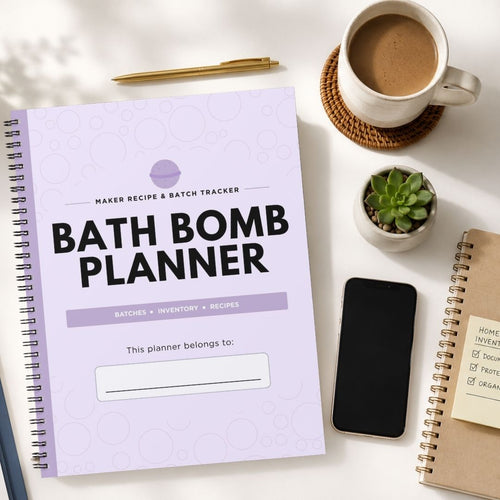 A spiral-bound Bath Bomb Planner notebook lies open on a white desk, surrounded by a coffee cup, pen, small succulent, smartphone, and another notebook. The planner cover is light lavender with black text reading 'BATH BOMB PLANNER' and a purple circular logo near the top.