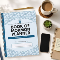 A spiral-bound Book of Mormon Planner with a light blue patterned cover is shown in a styled flat-lay on a white surface, surrounded by a coffee cup, a pen, a small succulent, a smartphone, and another notebook.