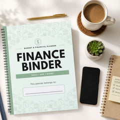 A spiral-bound 'Finance Binder' planner is displayed in a styled flat-lay arrangement on a white desk, surrounded by a cup of coffee, a smartphone, a small succulent, and other notebooks.