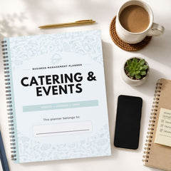 A spiral-bound business management planner titled 'Catering & Events' is shown in a styled flat-lay arrangement on a white surface, alongside a cup of coffee, a pen, a small succulent, a smartphone, and another notebook.