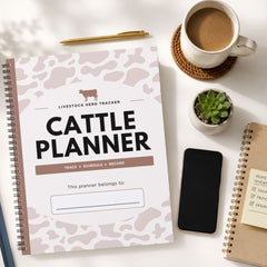 A spiral-bound Cattle Planner notebook with a white and dusty pink cow-print cover is displayed in a styled flat-lay on a white desk. The planner is surrounded by lifestyle props including a cup of coffee on a coaster, a small potted succulent, a smartphone, and another notebook.