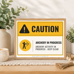 A framed novelty warning sign styled on a wooden table with plants and home decor. The sign features a yellow and white background with a black caution icon and the text 'CAUTION' and 'ARCHERY IN PROGRESS / ARCHERY ACTIVITY IN PROGRESS - KEEP CLEAR'.