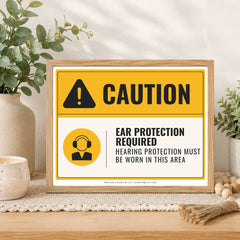 A framed caution sign with a yellow and white background, black text reading 'CAUTION' and 'EAR PROTECTION REQUIRED / HEARING PROTECTION MUST BE WORN IN THIS AREA,' styled on a tabletop with plants and home decor props.