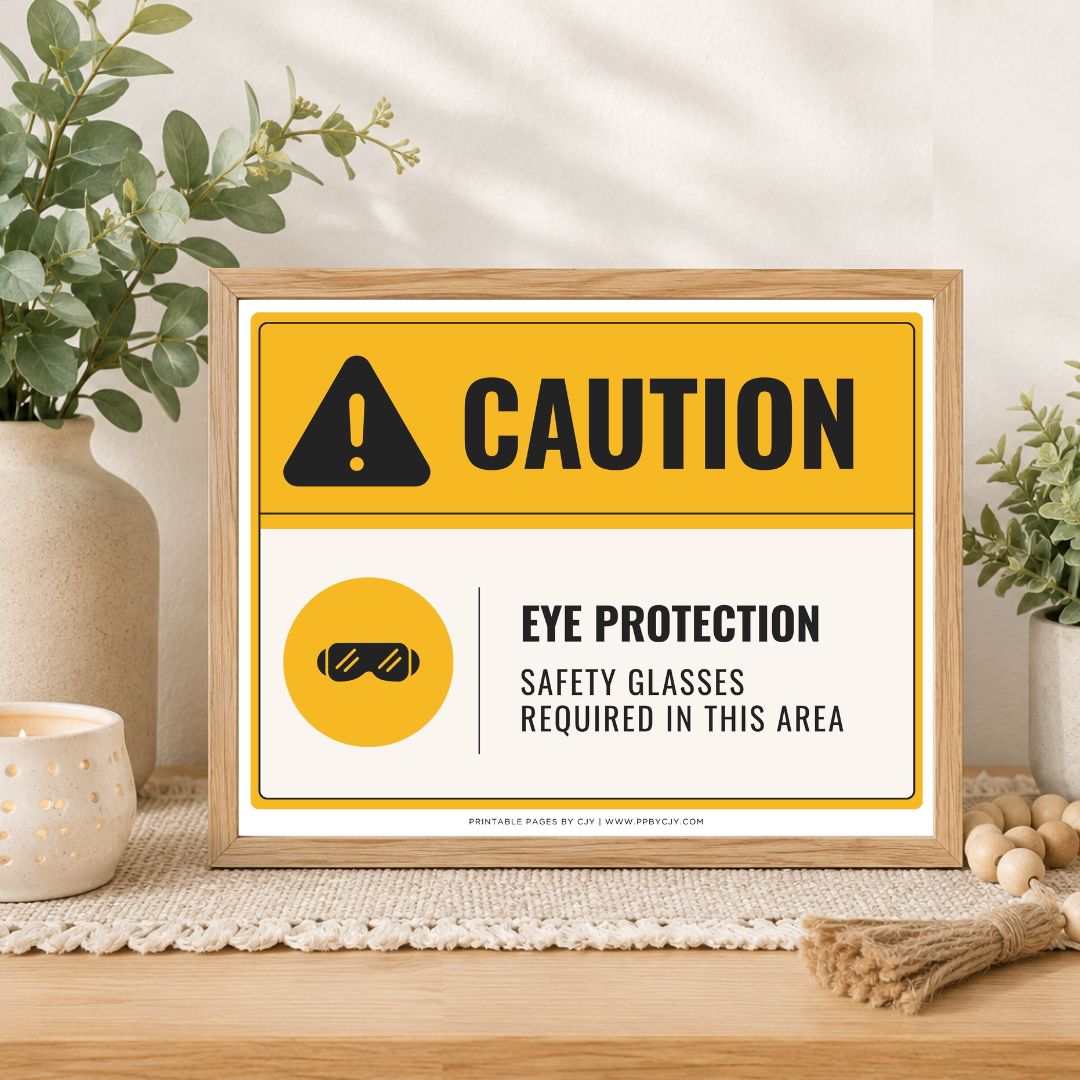 A framed caution sign on a wooden tabletop with plants and decor in the background. The sign reads “CAUTION” with an eye protection icon and text stating “EYE PROTECTION SAFETY GLASSES REQUIRED IN THIS AREA.