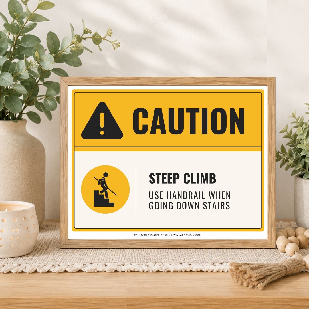 A framed caution-style sign on a tabletop in a styled interior setting. The sign has a yellow header with a black warning icon and the word CAUTION, plus a white lower section reading STEEP CLIMB and USE HANDRAIL WHEN GOING DOWN STAIRS.