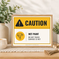 A framed caution sign with a yellow and white background, reading 'CAUTION' and 'WET PAINT DO NOT TOUCH SURFACE IS WET,' styled on a wooden surface with plants and home decor in the background.