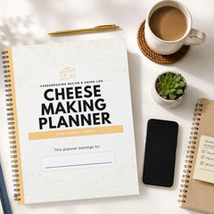 A spiral-bound 'Cheese Making Planner' lies on a white surface surrounded by lifestyle props, including a cup of coffee, a small succulent, a smartphone, and another notebook.