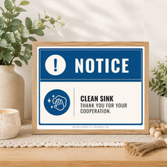 A framed novelty sink sign on a wooden table, styled with plants and a candle. The sign has a blue and white design with the text 'NOTICE' and 'CLEAN SINK THANK YOU FOR YOUR COOPERATION.