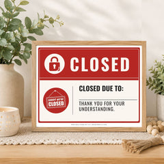 A framed 'Closed' sign with a red and white design sits on a wooden table in a styled home setting with plants and a candle.