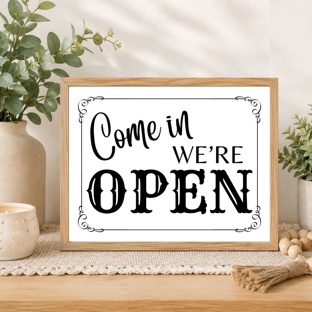 A framed decorative sign reading “Come in WE’RE OPEN” in black lettering on a white background, styled on a wooden surface with plants, a candle, and small wooden beads in a bright indoor setting.