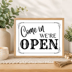 A framed decorative sign reading “Come in WE’RE OPEN” in black lettering on a white background, styled on a wooden surface with plants, a candle, and small wooden beads in a bright indoor setting.