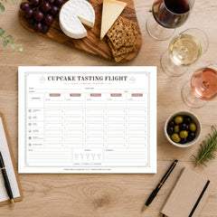 A printable 'cupcake Tasting Flight' chart laid on a wooden table, surrounded by food props like grapes, cheese, crackers, olives, and wine glasses. The chart is the product being shown, but the scene includes a real tabletop environment and styled props, so this fits Lifestyle rather than Product/Packaging.