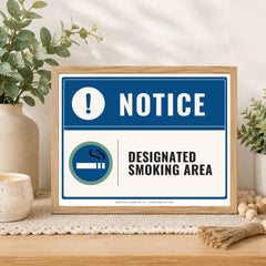 A framed smoking-area notice sign displayed on a wooden table in a styled home setting with plants and candles. The sign has a blue header with a white exclamation mark and the word 'NOTICE,' and a white lower section with the text 'DESIGNATED SMOKING AREA' and a blue-and-green cigarette icon.