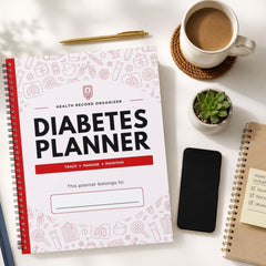A spiral-bound Diabetes Planner notebook is shown in a styled flat-lay arrangement on a white surface, surrounded by a coffee cup, a pen, a small succulent, a smartphone, and another notebook.