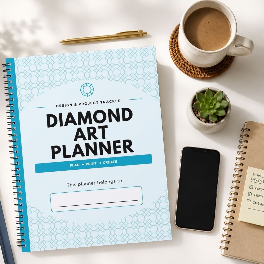 A spiral-bound 'Diamond Art Planner' with a light blue and white geometric pattern cover is displayed in a styled flat-lay arrangement on a white desk, surrounded by a cup of coffee, a smartphone, a small succulent, and other notebooks.