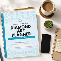 A spiral-bound 'Diamond Art Planner' with a light blue and white geometric pattern cover is displayed in a styled flat-lay arrangement on a white desk, surrounded by a cup of coffee, a smartphone, a small succulent, and other notebooks.