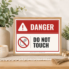 A framed warning sign on a tabletop in a styled home setting. The sign has a red and white background with the word 'DANGER' in white on red, a white warning triangle, a red prohibition symbol, and the words 'DO NOT TOUCH' in black.