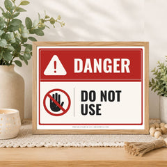 A framed warning sign with a red and white background, featuring the word 'DANGER' and a crossed-out hand symbol with the text 'DO NOT USE.' The sign is displayed on a tabletop in a styled interior setting with plants, a candle, and decorative props.