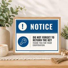 A framed printed notice sign on a wooden table, styled with plants, a candle, and decorative beads. The sign has a blue and white design with text reading 'NOTICE' and 'DO NOT FORGET TO RETURN THE KEY THANK YOU FOR YOUR COOPERATION.
