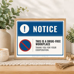 A framed novelty drug-free workplace sign displayed on a wooden table with decorative plants and a candle in the background.