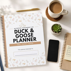 A spiral-bound planner titled 'Duck & Goose Planner' lies on a white surface, surrounded by a coffee cup, pen, small succulent, smartphone, and another notebook. The planner cover is white with a subtle duck and goose pattern and large black title text.