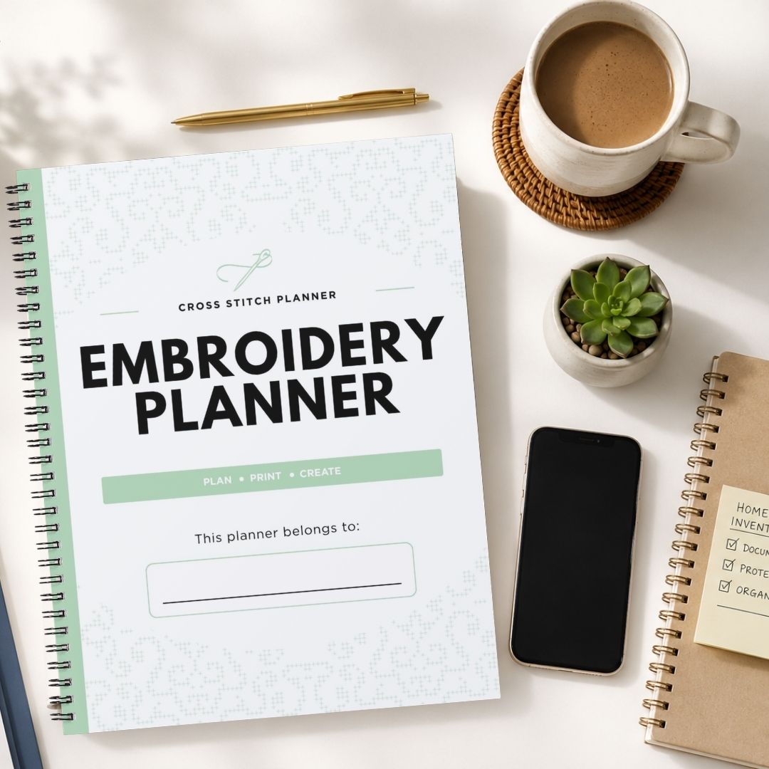 A spiral-bound embroidery planner with a white cover and mint green accents is displayed in a styled flat-lay arrangement on a white surface, surrounded by a cup of coffee, a pen, a small succulent, a smartphone, and another notebook.