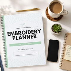 A spiral-bound embroidery planner with a white cover and mint green accents is displayed in a styled flat-lay arrangement on a white surface, surrounded by a cup of coffee, a pen, a small succulent, a smartphone, and another notebook.