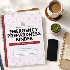 A spiral-bound emergency preparedness binder planner is shown in a styled flat-lay arrangement on a white desk, surrounded by a coffee cup, a pen, a small succulent, a smartphone, and another notebook.