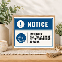 A framed hand-washing notice sign displayed on a wooden table in a styled home setting with plants and a candle. The sign features a blue and white design with the text 'NOTICE' and 'EMPLOYEES MUST WASH HANDS BEFORE RETURNING TO WORK'.