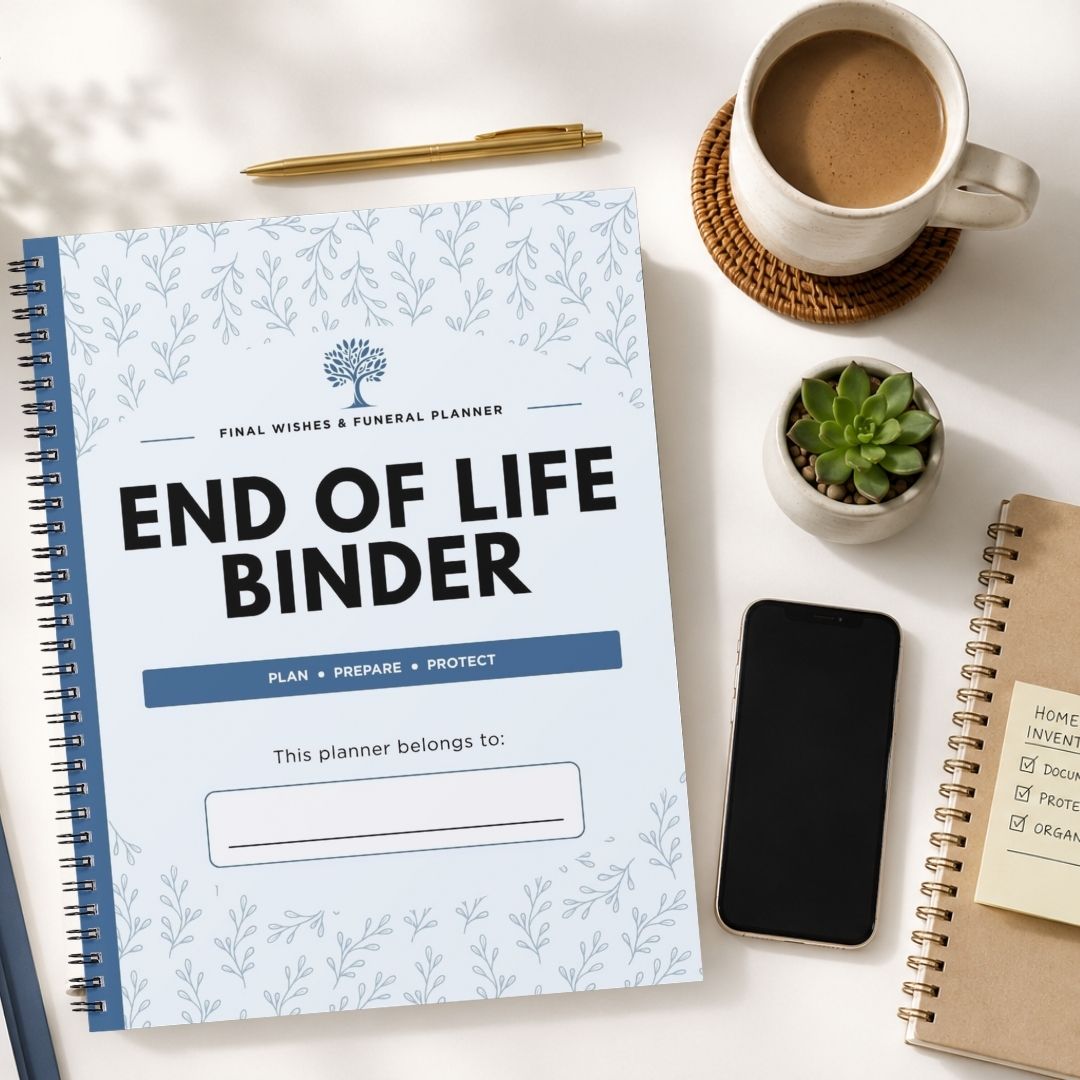 A spiral-bound 'End of Life Binder' planner is displayed in a styled flat-lay arrangement on a white desk alongside a cup of coffee, a smartphone, a small succulent, and other notebooks.