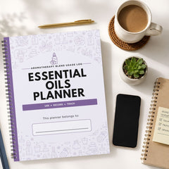 A spiral-bound 'Essential Oils Planner' with a white cover and purple accents, styled in a flat-lay arrangement on a white desk alongside a cup of coffee, a small succulent, a smartphone, and another notebook.