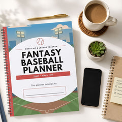 A spiral-bound fantasy baseball planner lies on a white desk surrounded by lifestyle props, including a cup of coffee, a small succulent, a smartphone, and another notebook.