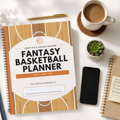 A spiral-bound fantasy basketball planner lies on a white desk surrounded by lifestyle props, including a cup of coffee, a small succulent, a smartphone, and another notebook.