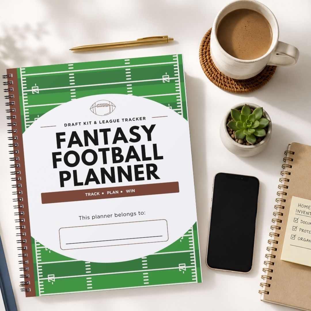 A spiral-bound fantasy football planner with a green football field design and large white title area, styled on a light tabletop with a coffee cup, pen, small plant, phone, and other notebooks around it.