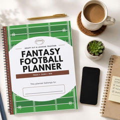 A spiral-bound fantasy football planner with a green football field design and large white title area, styled on a light tabletop with a coffee cup, pen, small plant, phone, and other notebooks around it.