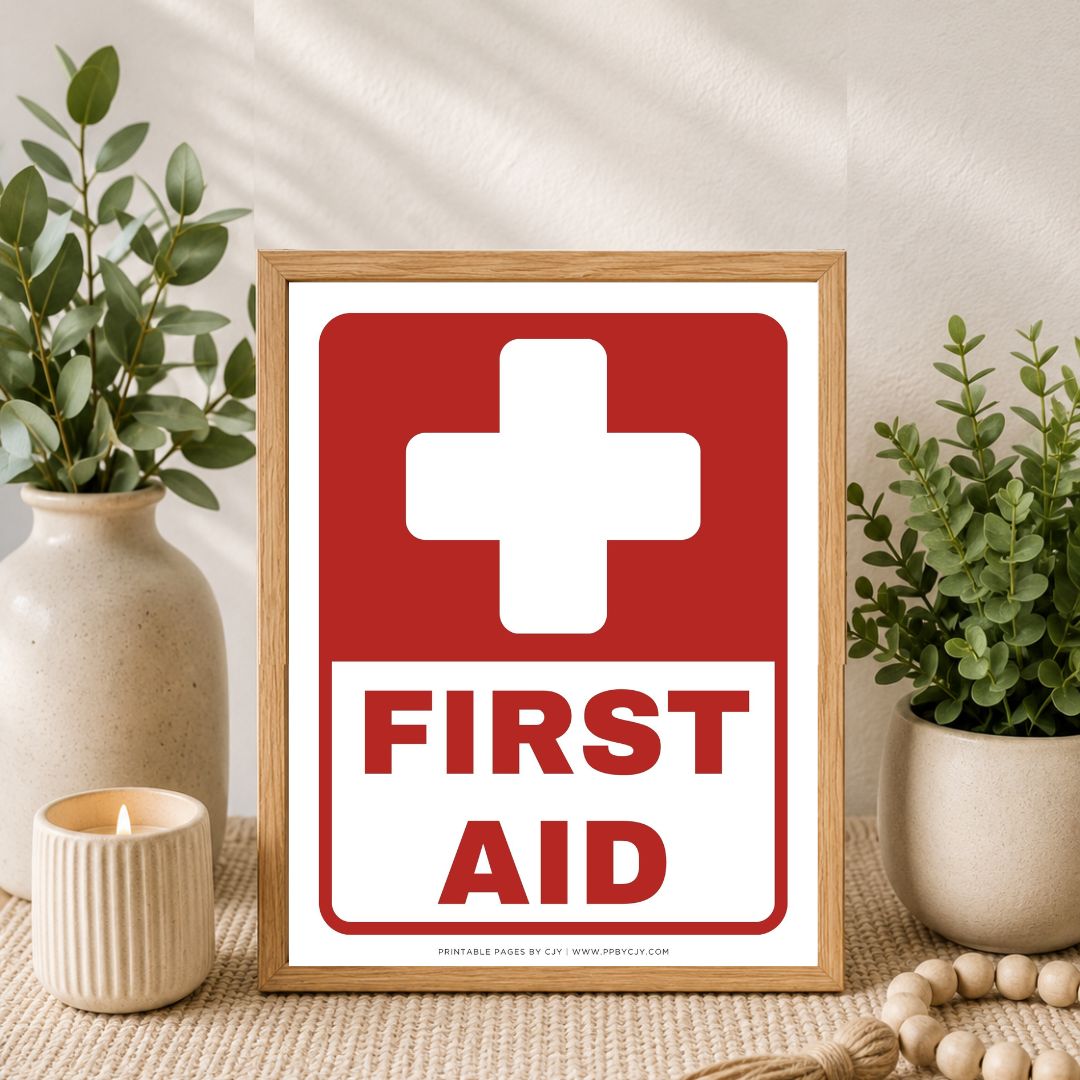 A framed first aid sign with a red cross and the words 'FIRST AID' in red on a white background, styled on a tabletop with potted plants, a candle, and a wooden bead runner.