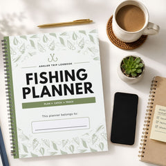 A spiral-bound fishing trip logbook titled 'FISHING PLANNER' is shown in a styled flat-lay arrangement on a white surface, alongside a cup of coffee, a small potted succulent, a smartphone, and another notebook.