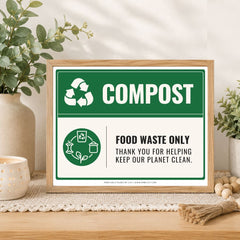 A framed compost sign displayed on a wooden table in a styled home setting. The sign features a green and white design with the word 'COMPOST' and icons indicating food waste only.