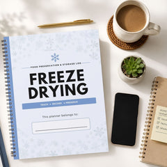 A spiral-bound freeze drying food preservation and storage log planner is shown in a styled flat-lay arrangement on a white surface, surrounded by a cup of coffee, a pen, a small succulent, a smartphone, and another notebook.