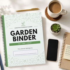 A spiral-bound garden planner titled 'GARDEN BINDER' lies on a white tabletop, surrounded by a coffee cup, a small succulent, a smartphone, and another notebook. The planner cover is white with light green botanical line drawings, a green band, and large black title text.