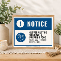 A framed printed notice sign on a tabletop in a styled interior setting. The sign has a blue border, a blue header with white 'NOTICE' text, and a white lower section with a blue hand icon and black text reading 'GLOVES MUST BE WORN WHEN PREPPING FOOD THANK YOU FOR YOUR COOPERATION.